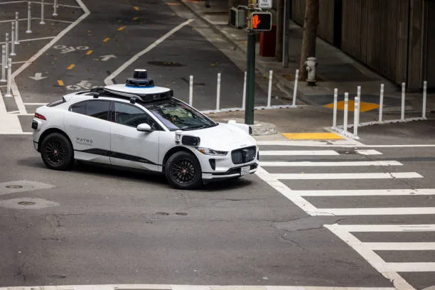 A Waymo autonomous vehicle, recognizable by its distinct sensor array, operates on public roads. Way
